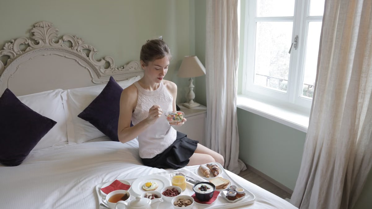 Calm female in elegant outfit sitting on bed and eating delicious breakfast served on tray in hotel room with classic interior in morning